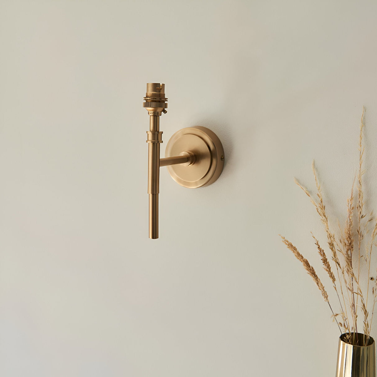 Endon Bevford Ikat wall light in aged brass with exposed B22 lampholder and rounded backplate, mounted on a pale wall beside dried grasses in a brass vase.