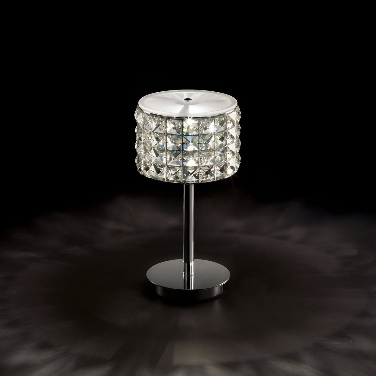 Introducing the Nebula Table Lamp in Chrome, a contemporary lighting marvel. It showcases a crystal-patterned cylindrical shade on an elegant metal stand, providing a gentle illumination against a dark backdrop.