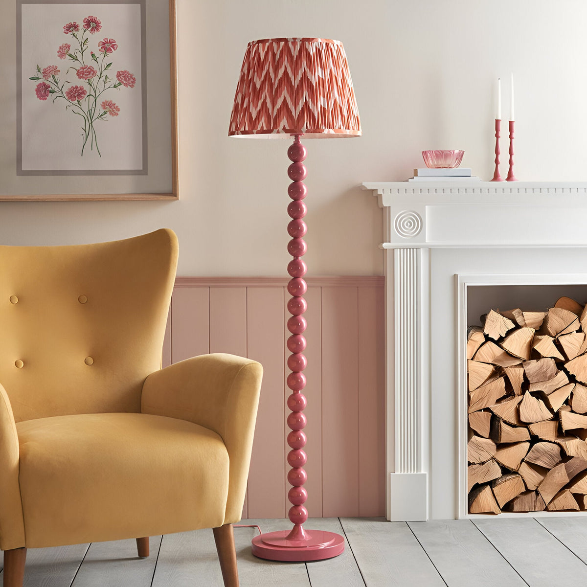 Brenleigh Zigzag Floor Lamp in pink, with a stacked spherical metal base and pleated apricot zigzag fabric shade, shown beside a mustard armchair and fireplace.