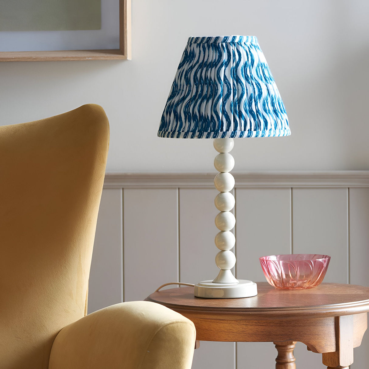 Endon Brenleigh Ripple table lamp with white stacked-sphere base and blue wave-print pleated shade, on a wooden side table beside a yellow armchair.