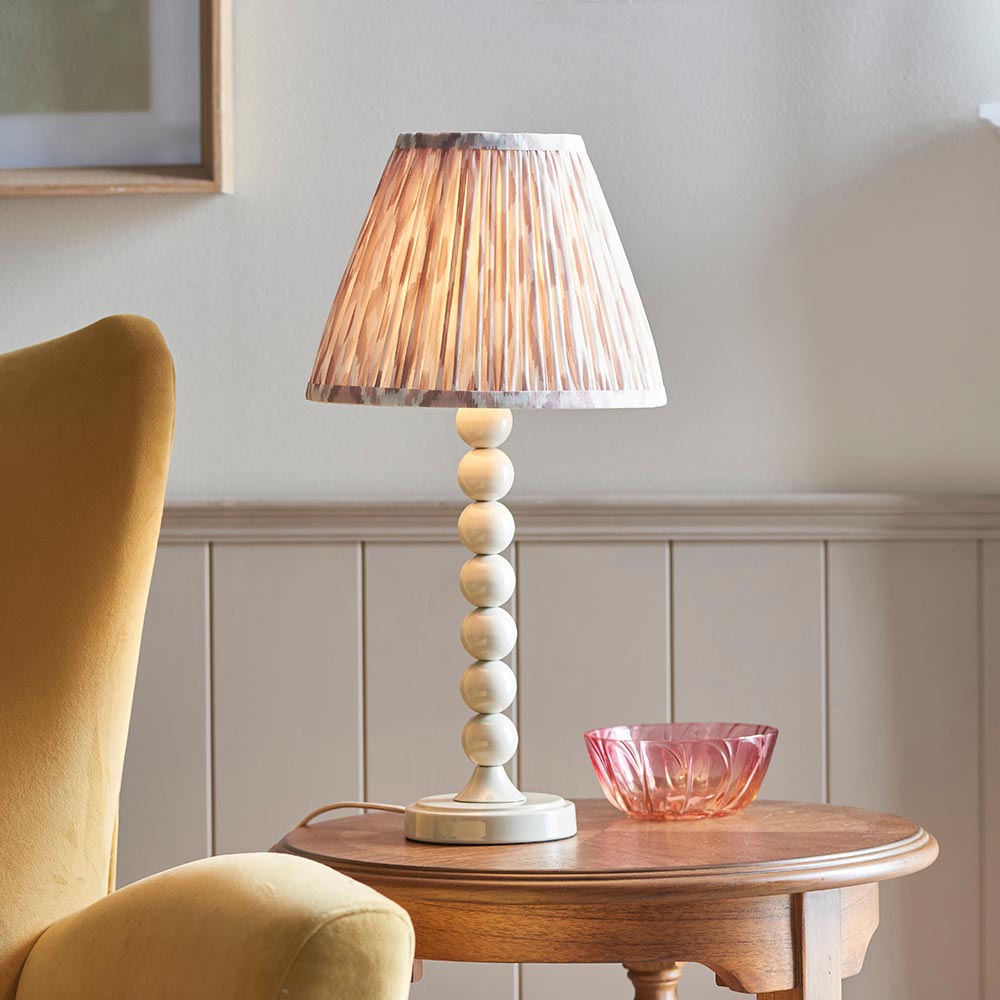 Brenleigh Ikat table lamp with white stacked-sphere base and pleated natural fabric shade, shown on a wooden side table beside a pink glass bowl.