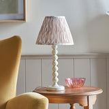 Brenleigh Ikat Table Lamp with stacked-sphere white base and pleated pink-toned Ikat shade, shown on a wooden side table beside a pink glass bowl.