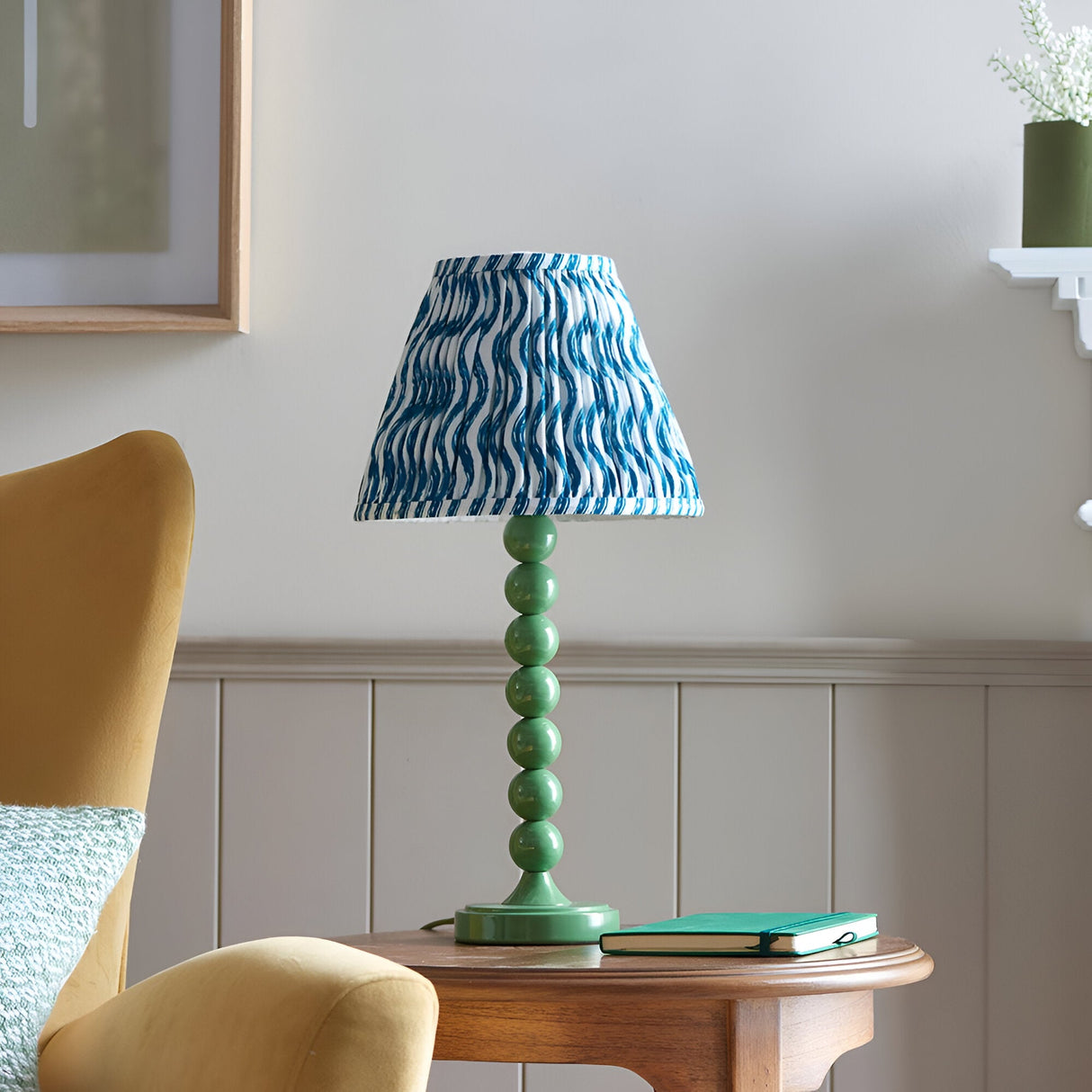Endon Brenleigh Ripple table lamp with green stacked-sphere metal base and blue wave-print pleated shade on a wooden side table beside a mustard chair.