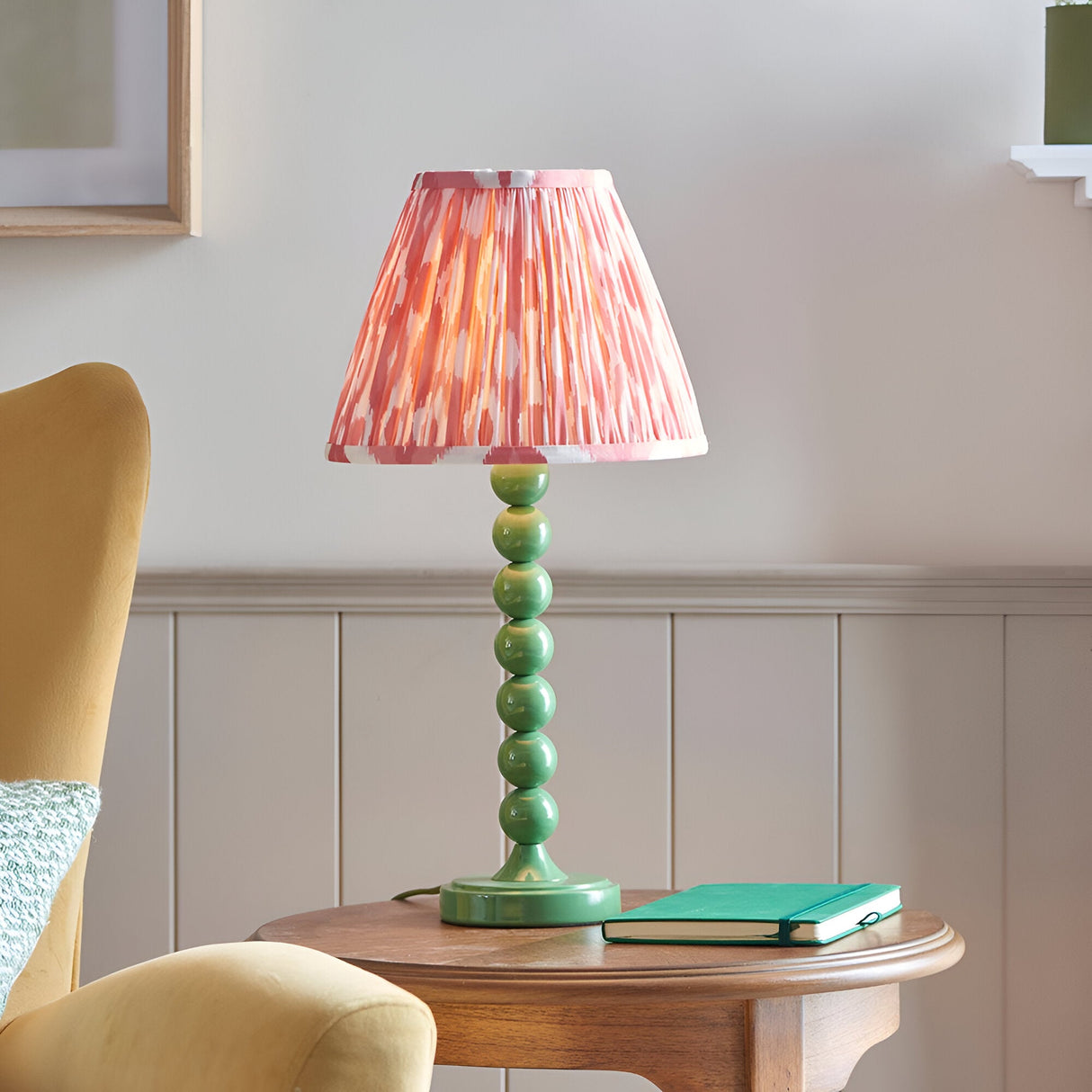 Endon Brenleigh Ikat table lamp with a stacked green sphere base and pink pleated Ikat shade on a wooden side table beside a yellow armchair.