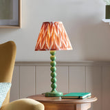 Brenleigh Zigzag Table Lamp in green, with a stacked-sphere metal base and pleated orange zigzag shade, shown on a side table beside a notebook.