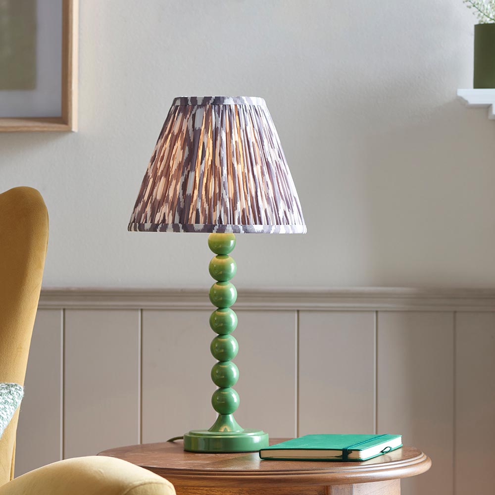 Brenleigh Ikat Table Lamp with green stacked-sphere metal base and grey ikat-print pleated fabric shade, shown on a side table in a living room.