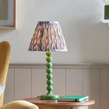 Brenleigh Ikat Table Lamp with green stacked-sphere metal base and grey ikat-print pleated fabric shade, shown on a side table in a living room.