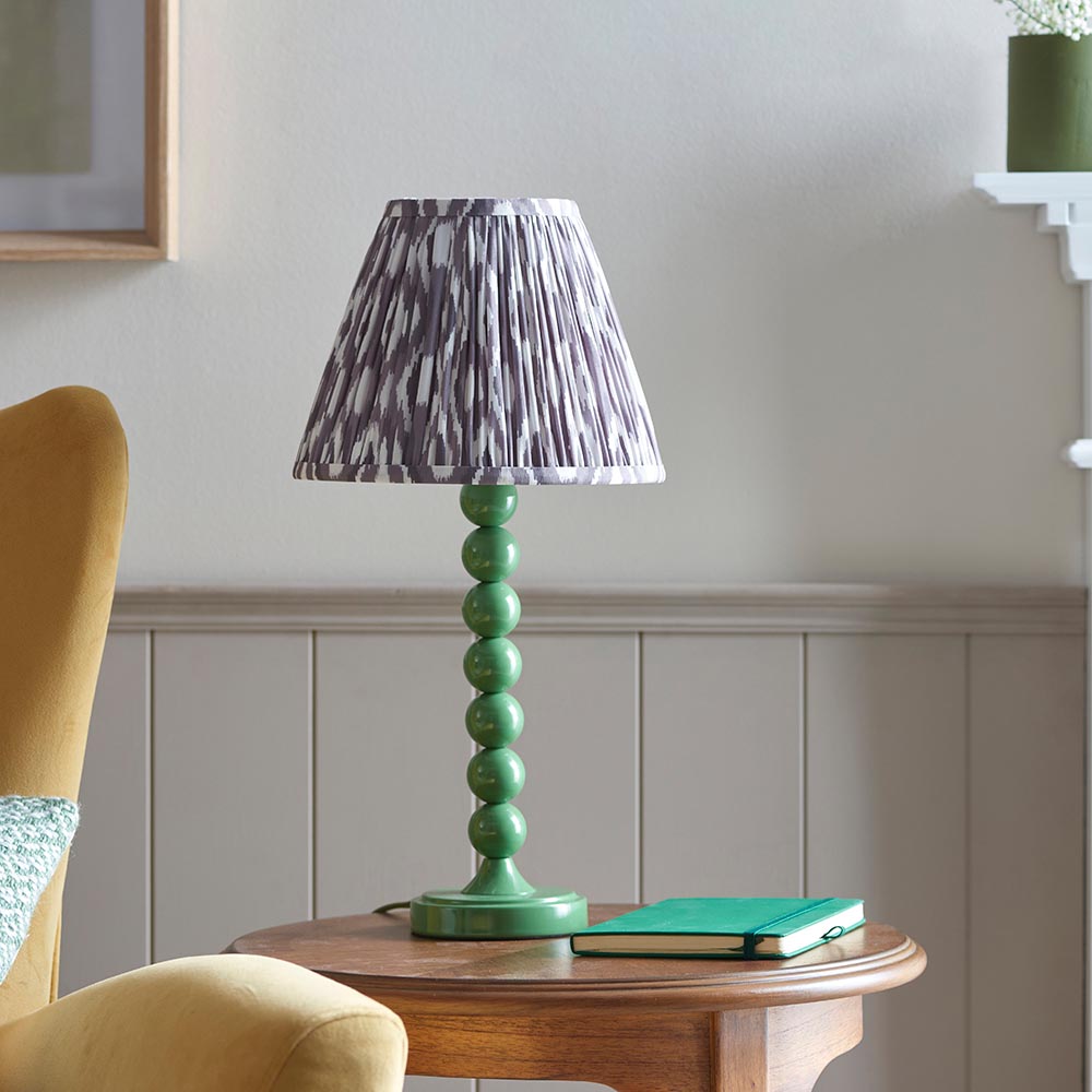 Brenleigh Ikat Table Lamp with green stacked-sphere gloss base and pleated grey Ikat fabric shade, shown on a side table in a living room.