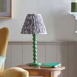 Brenleigh Ikat Table Lamp with green stacked-sphere gloss base and pleated grey Ikat fabric shade, shown on a side table in a living room.
