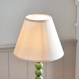 Brenleigh Ivy Table Lamp with white cotton-mix Ivy shade and green stacked-sphere metal base, shown against a light grey wall setting.