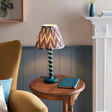 Endon Brenleigh Zigzag table lamp with teal stacked-sphere metal base and pleated grey zigzag shade on a wooden side table beside a mustard armchair.
