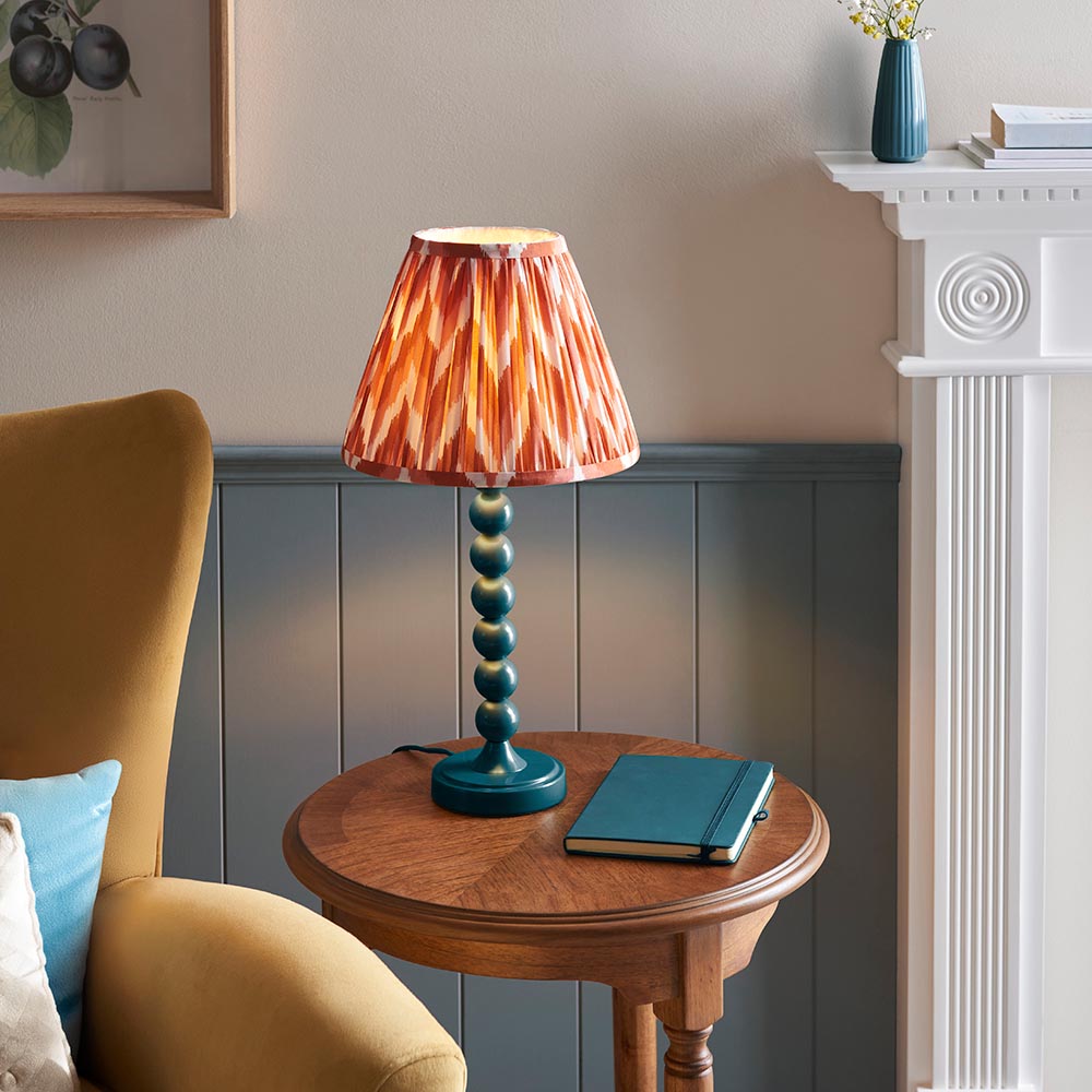Brenleigh Zigzag Table Lamp with orange pleated Zigzag shade and blue stacked-sphere metal base, styled on a wooden side table beside a notebook.