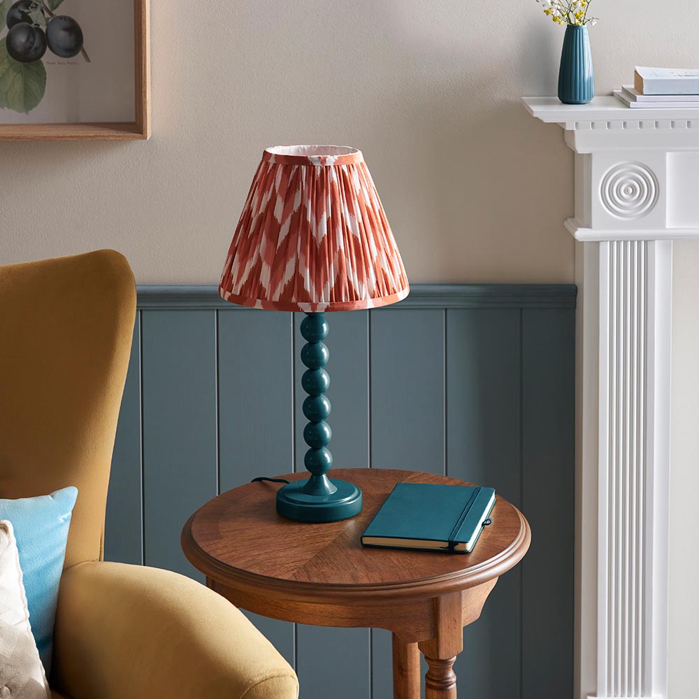 Brenleigh Zigzag Table Lamp with teal stacked-sphere metal base and pleated red-orange zigzag shade, styled on a wooden side table beside a mustard armchair.