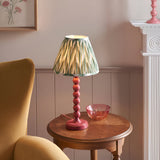 Endon Brenleigh Zigzag table lamp with pink stacked-sphere base and green pleated zigzag shade on a wooden side table beside a mustard armchair.