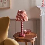 Brenleigh Zigzag Table Lamp in pink, stacked-sphere metal base with pleated zigzag-pattern shade, shown on a side table beside a glass bowl.