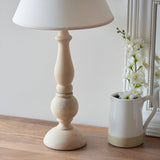 Brenhurst Table Lamp in vintage white, hand-turned solid wood base with tapered cotton mix shade, shown beside a jug of white flowers on a wooden surface.