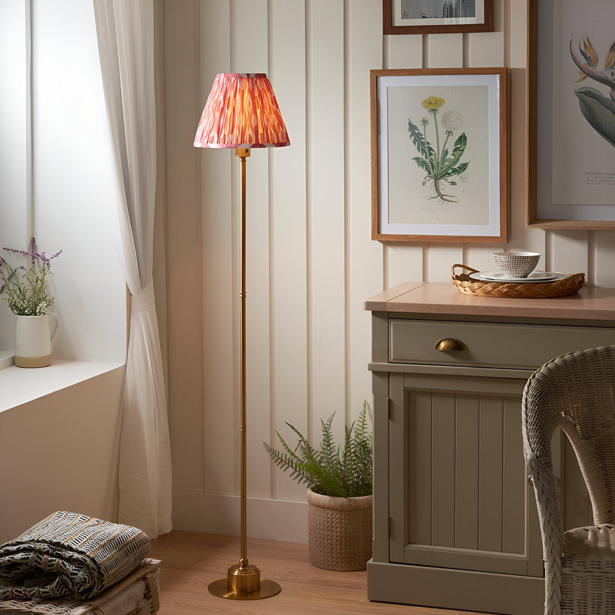 Endon Ashstead LED floor lamp with brushed brass stem and coral pink Ikat pleated shade, shown in a cottage-style room beside a cabinet and window.