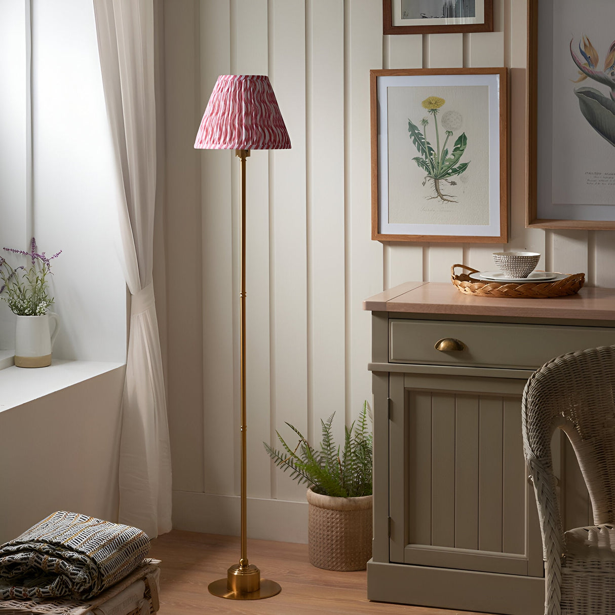 Endon Ashstead LED floor lamp with brushed brass stem and pink and white ripple pleated shade in a cottage room beside a wooden cabinet and botanical prints.