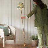 Woman in green linen adjusts the Endon Ashstead LED floor lamp with brushed brass stem and green zigzag pleated shade beside a wicker chair.