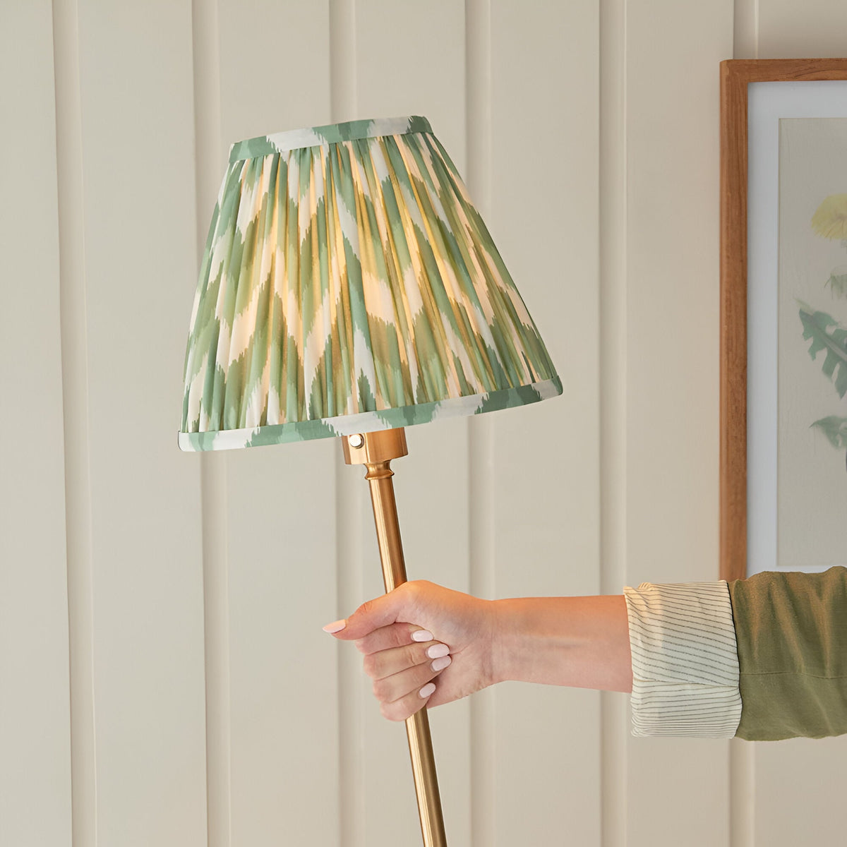 Hand holding the brass stem of the Endon Ashstead LED floor lamp, topped with a green and white zigzag pleated shade against panelled wall.