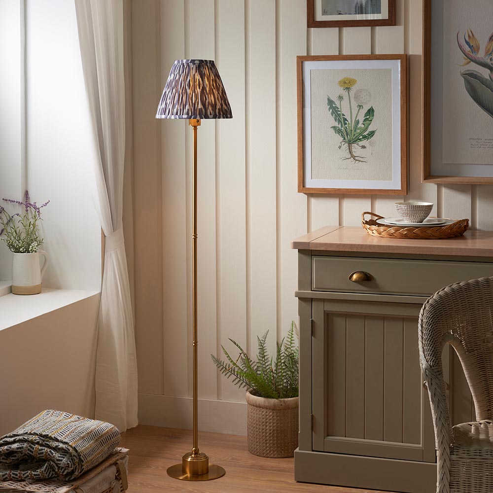 Endon Ashstead LED floor lamp with a brushed brass stem and grey Ikat pleated shade, styled in a bedroom beside botanical prints and a green cabinet.