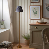 Endon Ashstead LED floor lamp with brushed brass stem and grey Ikat pleated shade, beside a cream cabinet, framed botanical prints and fern in a living space.