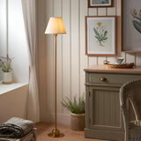 Endon Ashstead LED floor lamp with brushed aged brass stem and vintage white Ivy fabric shade, lit beside a cream panelled wall in a cosy living room.