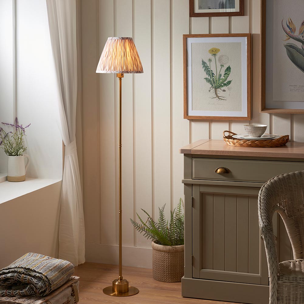 Endon Ashstead LED floor lamp with brushed brass stem and natural pleated Ikat shade, lit beside a cream cabinet and botanical prints in a cosy room.
