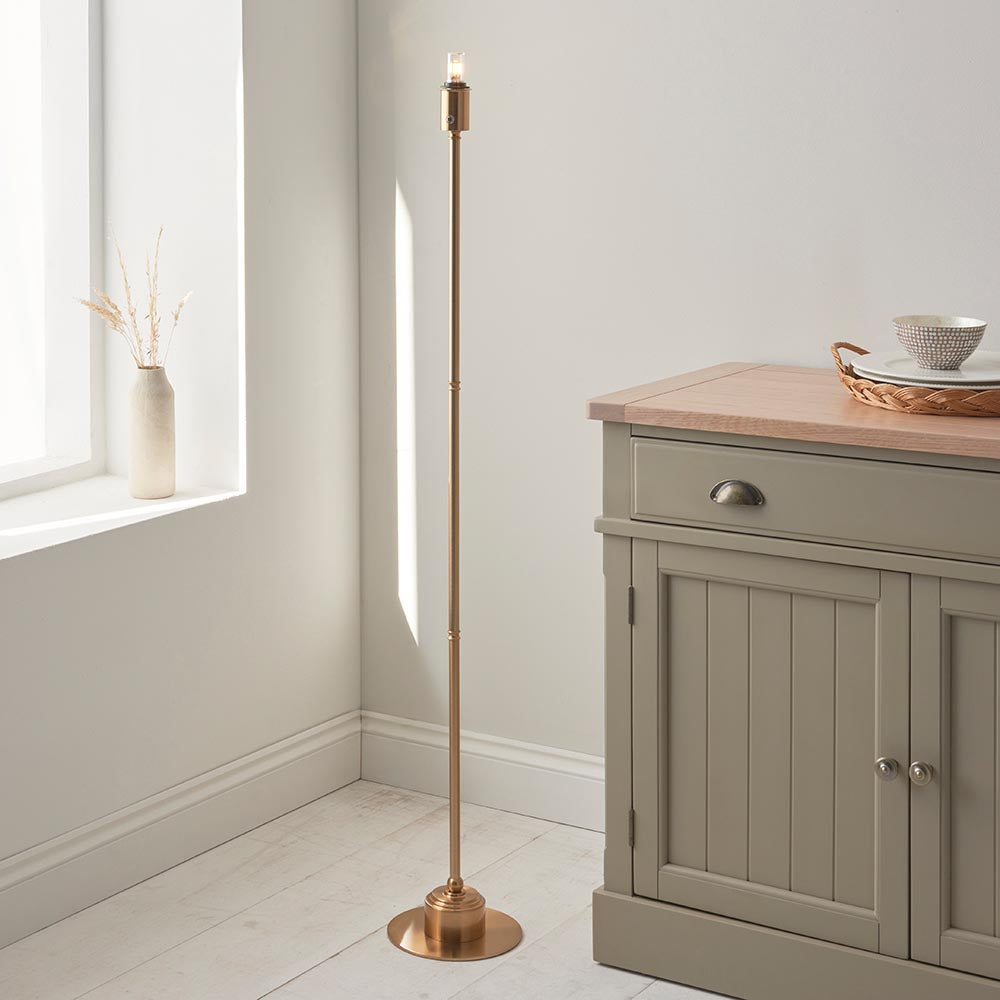 Endon Ashstead LED floor lamp on a slim brushed brass stem with round base and small bulb top, beside a sage sideboard and light window nook.