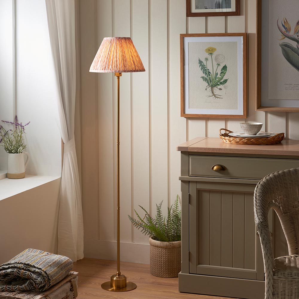 Ashstead LED Leaf Floor Lamp with pleated fabric shade on a slim brushed brass stem, shown in a living room beside a wooden cabinet.