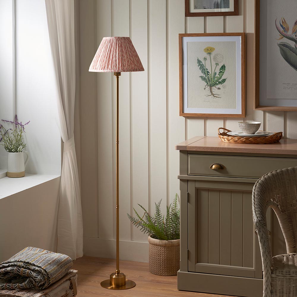 Ashstead LED Leaf Floor Lamp with brushed brass stem and pleated pink fabric shade, shown in a living room beside a cabinet and framed prints.
