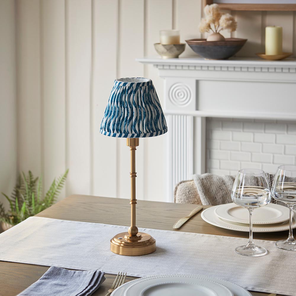 Ashstead LED Ripple Table Lamp with brass stand and blue-and-white patterned fabric shade, shown on a dining table in a living room setting.