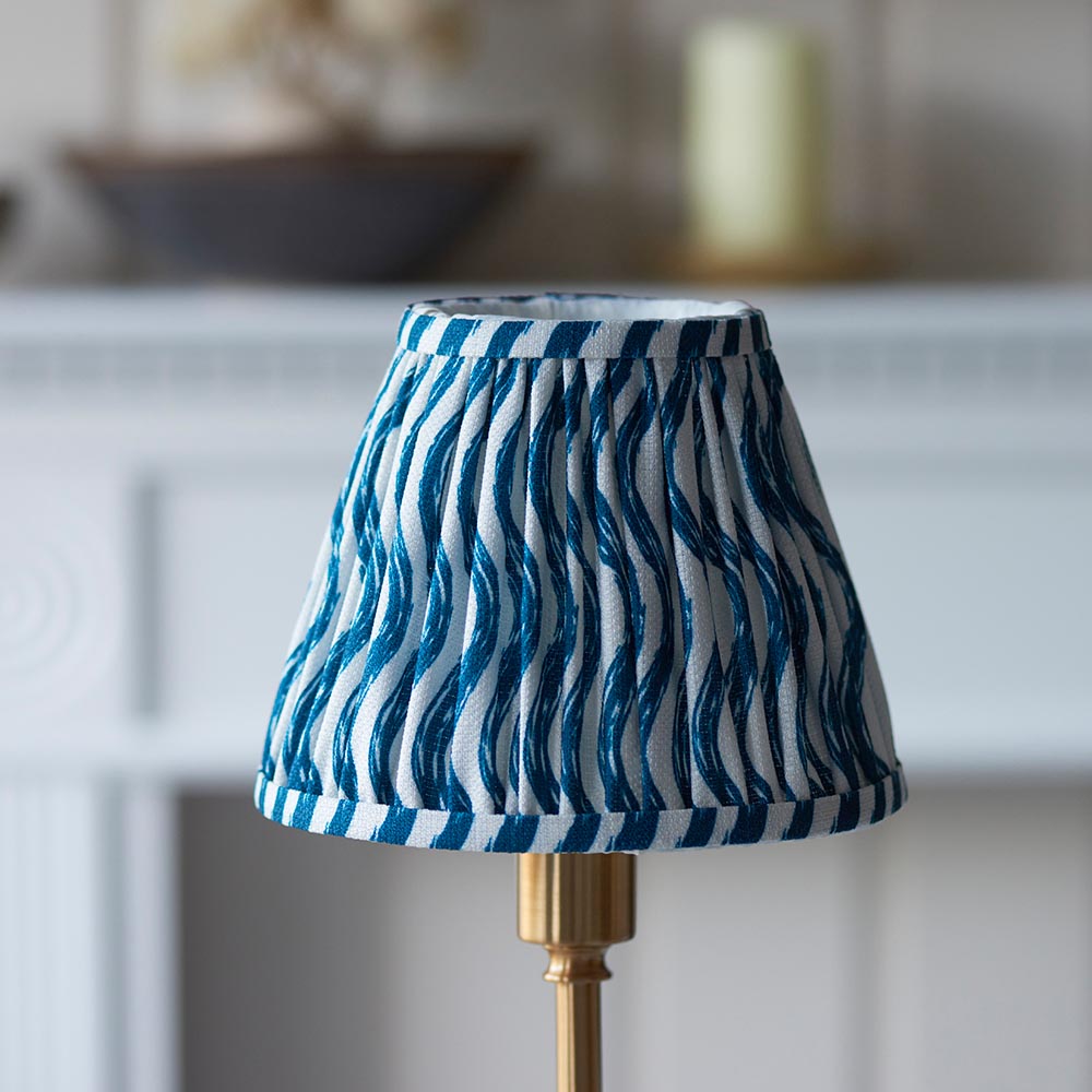 Ashstead LED Ripple Table Lamp with pleated blue and white striped fabric shade on a brass base, shown on a mantelpiece in a living room setting.