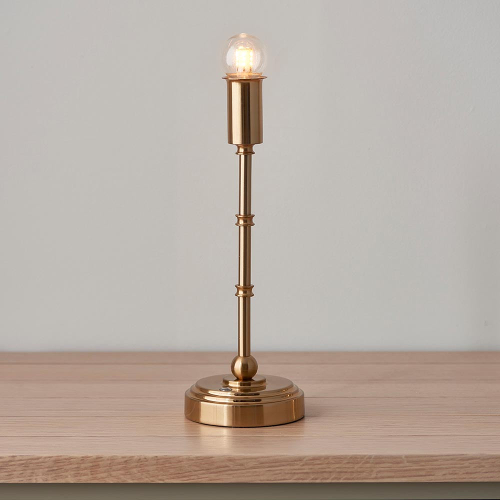 Ashstead LED table lamp with slim brass stem, round base, and exposed warm-white bulb, shown on a wooden surface against a plain background.