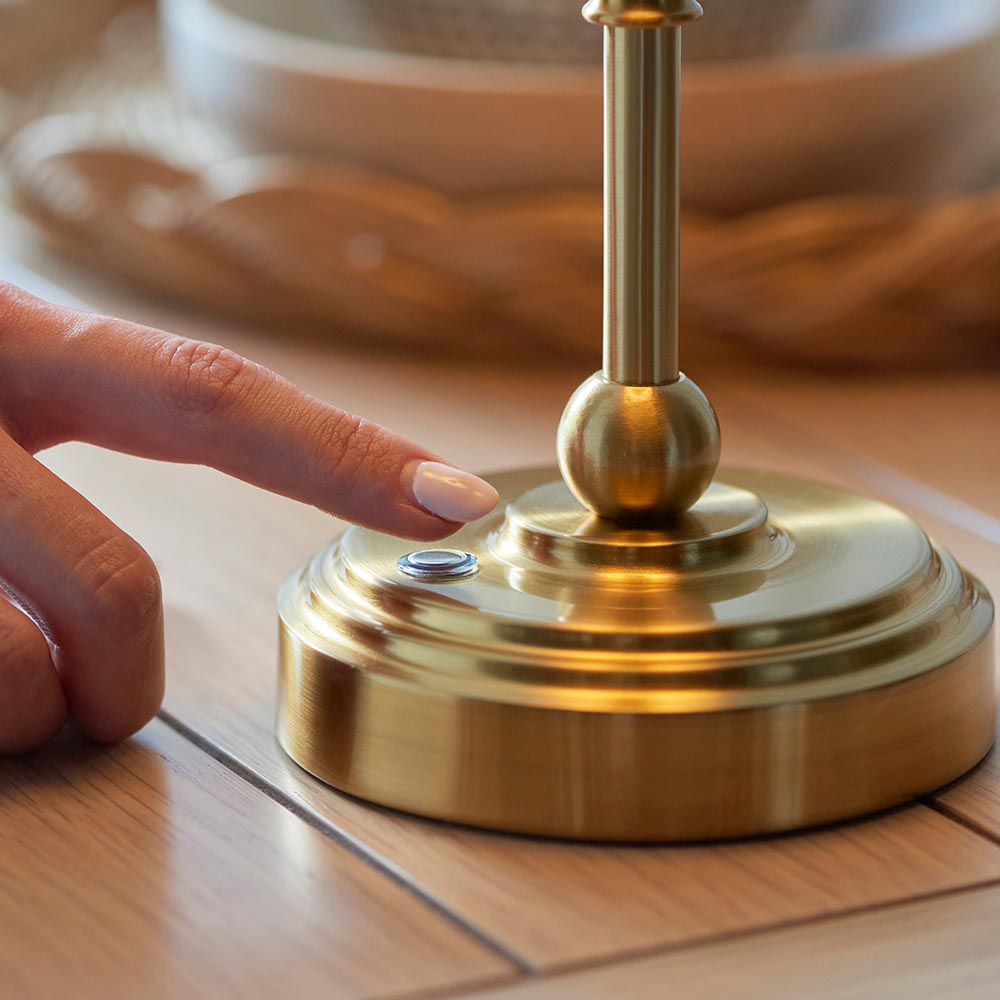 Ashstead LED Ripple Table Lamp base in brushed brass with touch dimmer button, shown close-up as a finger adjusts the control on a wooden surface.