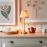 Endon Ashstead LED table lamp with brushed brass stem and pleated pink ikat shade, lit on a wood counter beside botanical prints, a red teapot and dishes.
