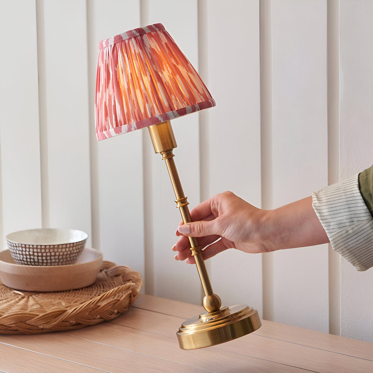 Hand adjusting the Endon Ashstead cordless LED table lamp with brushed brass stem and pleated pink Ikat shade on a wooden sideboard.