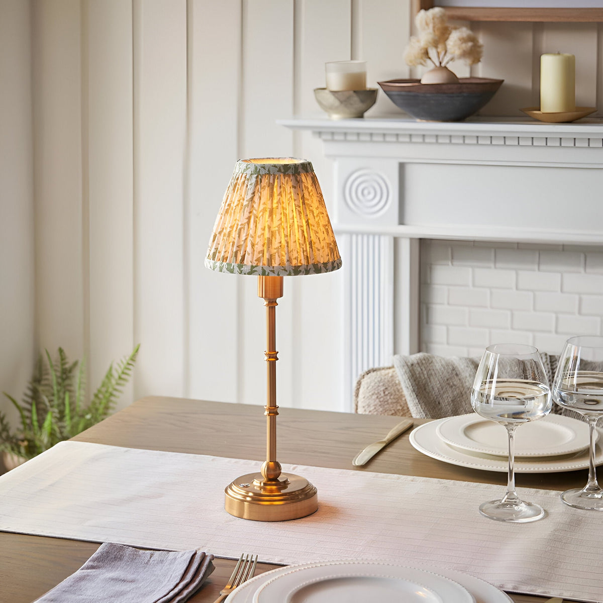 Ashstead LED Leaf Table Lamp with brass base and stem, featuring a pleated white and green leaf-print fabric shade, shown lit on a dining table.