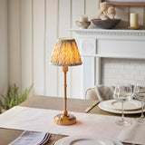 Ashstead LED Leaf Table Lamp with brass base and stem, featuring a pleated white and green leaf-print fabric shade, shown lit on a dining table.