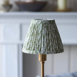 Ashstead LED Leaf Table Lamp with pleated white and green leaf-print fabric shade on a brass base, shown on a mantelpiece in a light room setting.