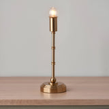 Ashstead LED table lamp with brass candlestick base and stem, lit exposed globe bulb, on a wooden surface against a plain grey background.
