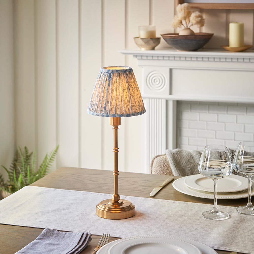 Ashstead LED Leaf Table Lamp with brass base and pleated blue and white leaf-print shade, shown on a dining table beside glassware and a white mantelpiece.