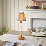 Ashstead LED Leaf Table Lamp with brass base and pleated blue and white leaf-print shade, shown on a dining table beside glassware and a white mantelpiece.