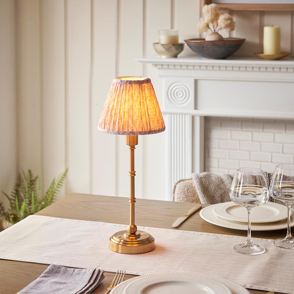 Endon Ashstead LED table lamp with brushed brass stem and pleated peach leaf-print shade on a dining table set with wine glasses near a white fireplace.