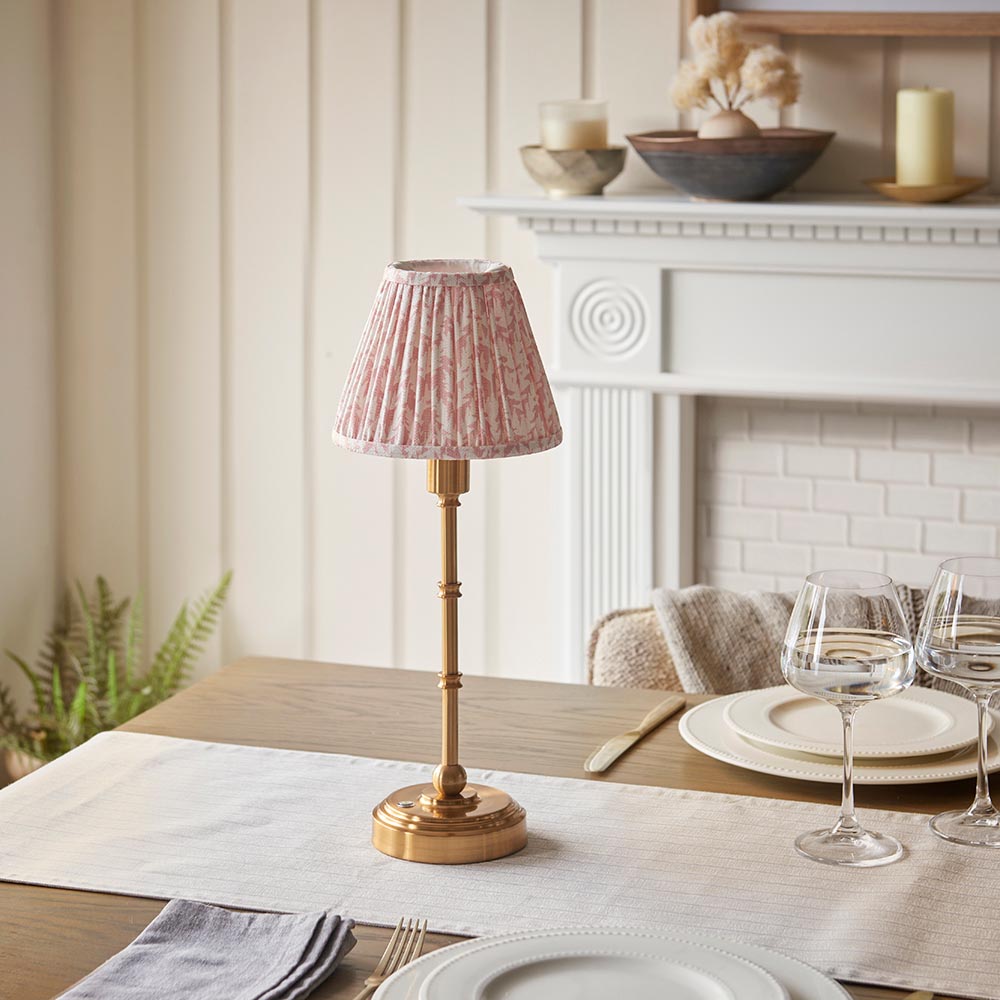 Endon Ashstead LED table lamp with brushed brass stem and pleated pink leaf-print shade, on a dining table set with glassware near a white fireplace.
