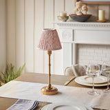Endon Ashstead LED table lamp with brushed brass stem and pleated pink leaf-print shade, on a dining table set with glassware near a white fireplace.