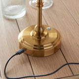 Close-up of the Endon Ashstead LED table lamp's brushed aged brass base on a wooden table, showing the USB-C charging port with cable connected.