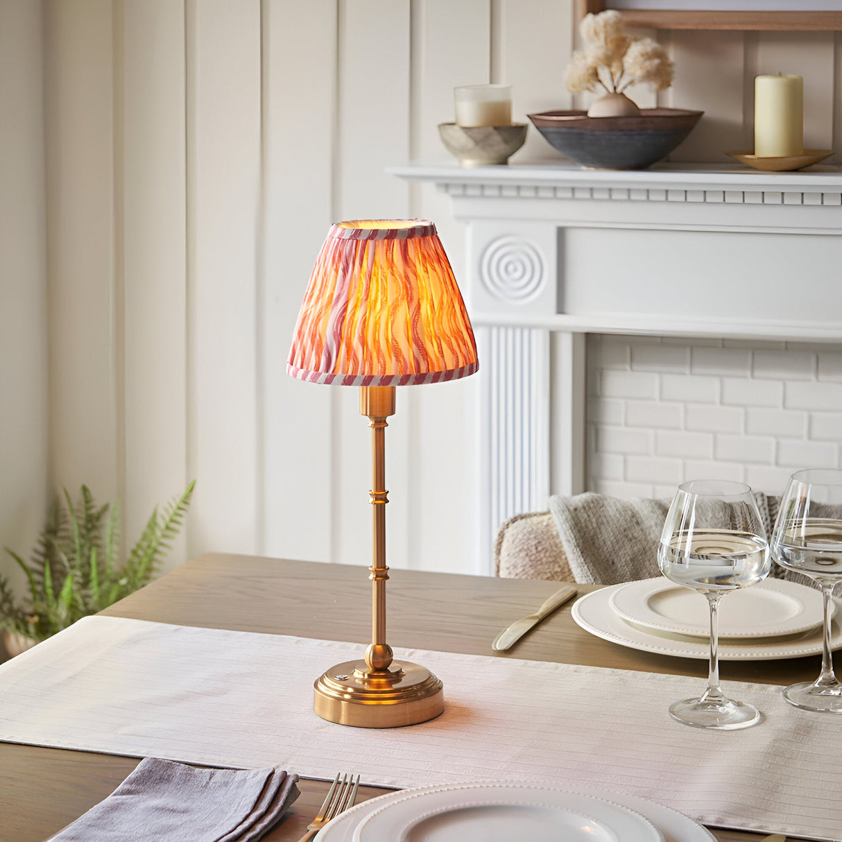 Endon Ashstead LED table lamp with brushed brass stem and pleated pink ripple-print shade, styled on a dining table beside a white fireplace.