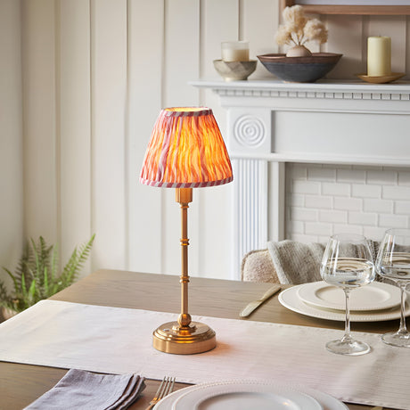 Endon Ashstead LED table lamp with brushed brass stem and pleated pink ripple-print shade, styled on a dining table beside a white fireplace.