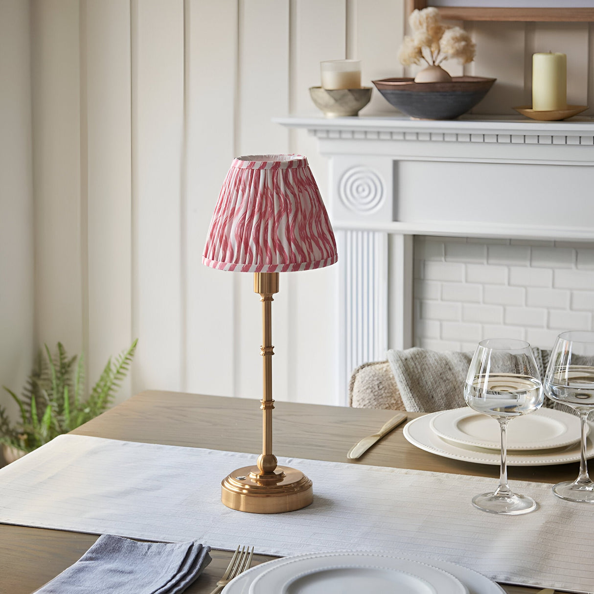 Endon Ashstead LED Ripple cordless table lamp with brushed brass stem and pleated pink and white rippled shade on a dining table by a white fireplace.